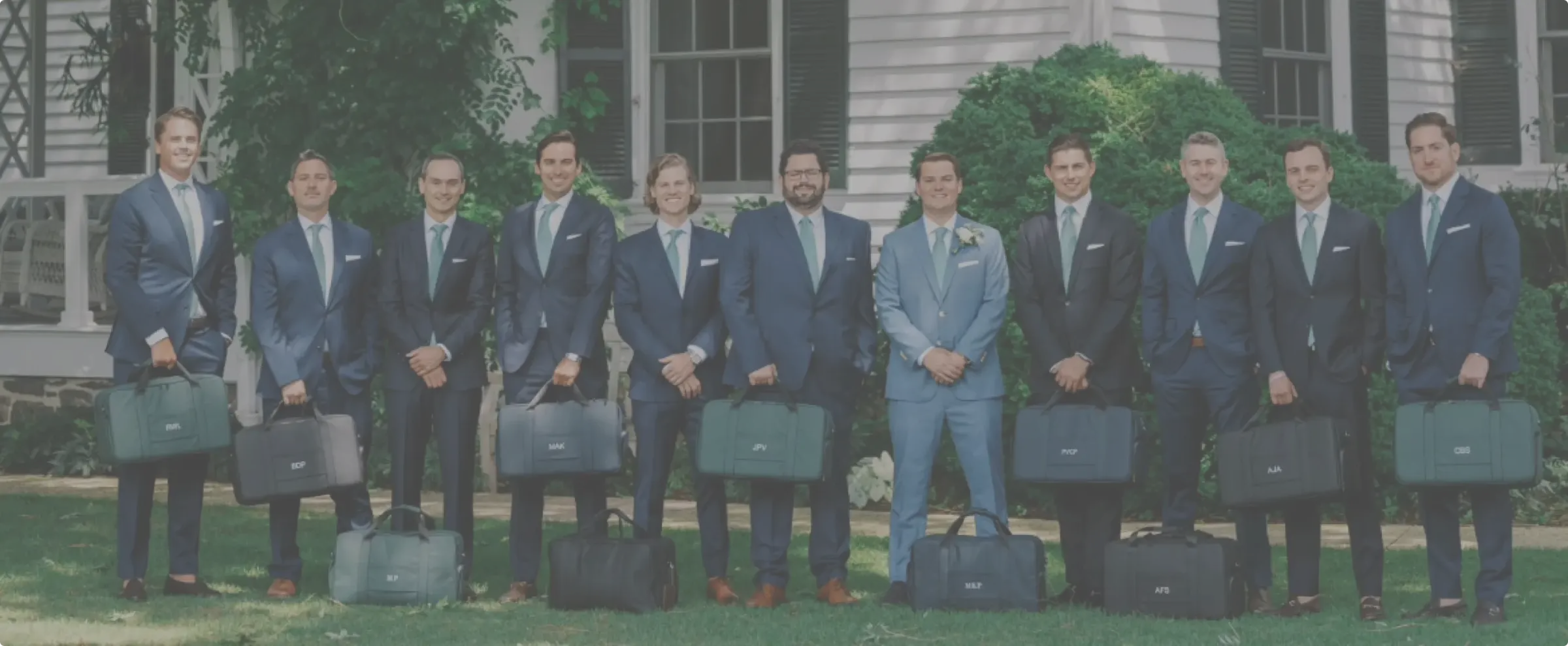 Groomsmen in matching suits holding monogrammed duffel bags on a lawn outside a house.