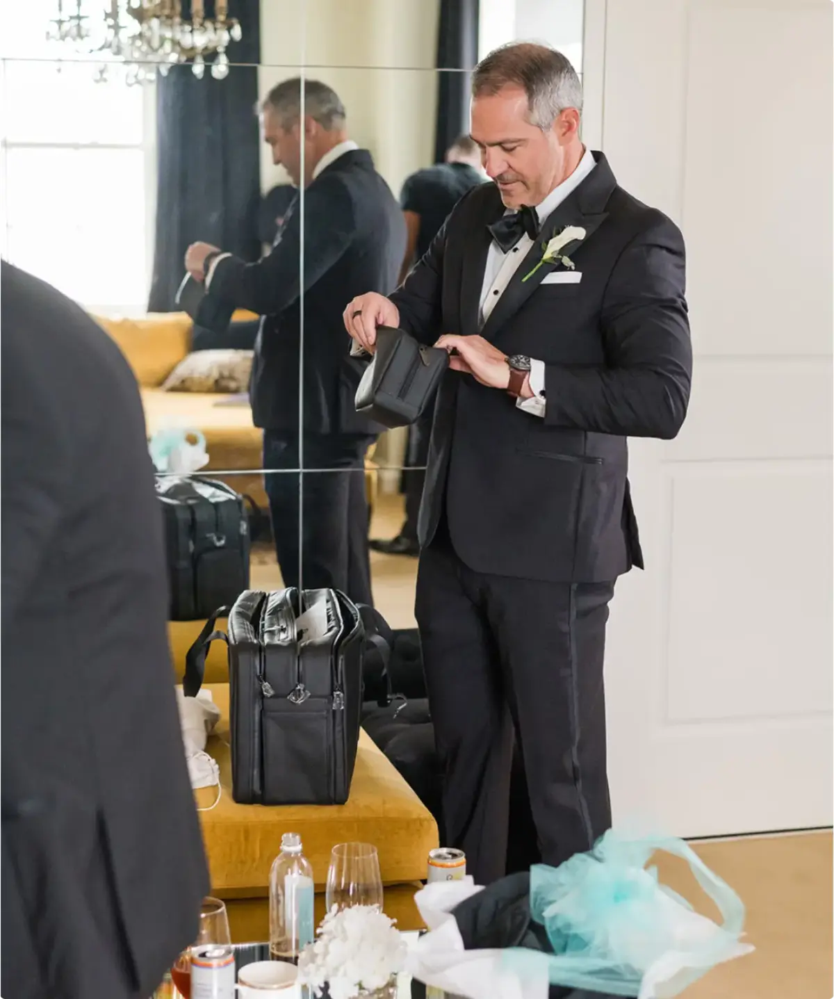 Groom in a tuxedo opening a leather kit beside a black garment duffel bag.